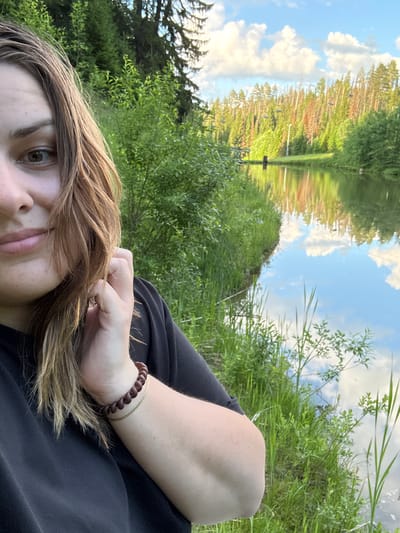 A young woman with light brown hair is taking a selfie outdoors, with a body of water and a forested area in the background. The location is identified as Nechkino, Russia. She is wearing a black t-shirt and a brown beaded bracelet on her right wrist, with a thin gold chain beneath it. Her hand is raised to her face, touching her hair. The water behind her reflects the trees, sky, and clouds, creating a serene natural scene. The trees appear to be a mix of evergreen and deciduous, with some showing signs of seasonal change. The sky is blue with scattered white clouds.