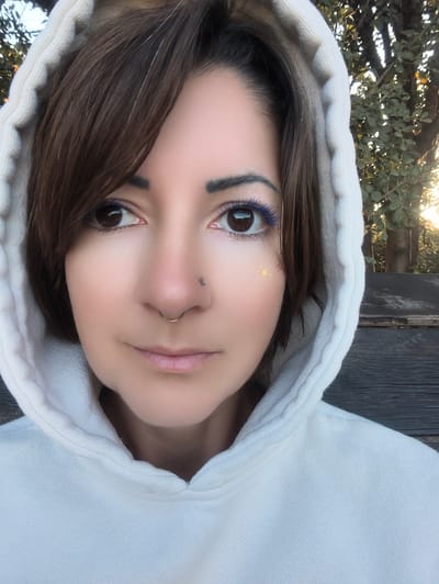 A close-up, eye-level portrait features an individual, likely female, wearing a light-colored (off-white) hooded sweatshirt with the hood pulled up around their head. The person has brown hair with a fringe, brown eyes, and a neutral expression, gazing directly forward. Both lower eyelids are accented with vibrant blue eyeliner. A small cluster of yellow and pink glitter specks is visible on the cheek beneath the right eye. The left nostril is adorned with a small silver stud piercing, and a gold-toned hoop piercing is present in the septum. The background is out of focus, revealing green foliage from trees or bushes on the right side and a dark, weathered wooden surface (possibly a bench or fence) behind and to the left. Soft, warm natural light illuminates the scene, appearing to originate from the right rear, filtering through the foliage. The location details in the city of Shaalvim, Israel are not discernible from the visual information provided in the image.