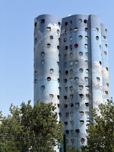 The image shows the exterior of the "Tours Nuages" (Cloud Towers) building in Nanterre, France, designed by architect Renzo Piano. The building is characterized by its distinctive cylindrical towers, clad in a mosaic of light blue tiles, with numerous circular and irregularly shaped windows. Some windows are open, revealing interior elements. Trees with green foliage are visible in the foreground, partially obscuring the lower levels of the building, and a wire fence runs across the bottom of the frame. The sky is clear and blue. There are no people visible in the image.