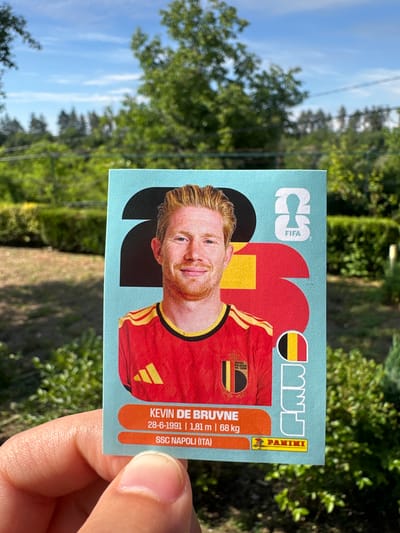 A vertical medium shot shows a hand holding a Panini soccer sticker of Kevin De Bruyne. The sticker is blue with red, yellow, and black graphic elements. De Bruyne, with his signature blonde hair and beard, is depicted in a red soccer jersey with yellow trim. Text on the sticker includes "KEVIN DE BRUYNE," his birthdate "28-6-1991," height "1,81 m," weight "68 kg," and club "SSC NAPOLI (ITA)." The FIFA and Panini logos are also visible. The background is a soft-focus view of green trees and a blue sky with white clouds, suggesting an outdoor setting on a bright, possibly sunny day. The hand holding the sticker is a pale complexion with visible fingernails and fingerprints. The overall impression is that of someone showing off a collectible item outdoors.