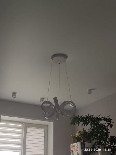 A modern, white, abstract ceiling light fixture is centrally located in a domestic setting. The fixture consists of two looped, metallic-like elements suspended from the ceiling by thin wires. The room appears to be illuminated by natural light from a window with horizontal blinds on the left side of the image. Two smaller, cylindrical white lights are also visible on the ceiling, one to the left and one to the right of the main fixture. A large, leafy green plant is situated on the right side of the image, partially obscuring the background. A date and time stamp, "23.06.2026 12:28", is present in the bottom right corner. The overall impression is that of a well-lit interior space with contemporary decor. The location is identified as Kuyuki, Russia.