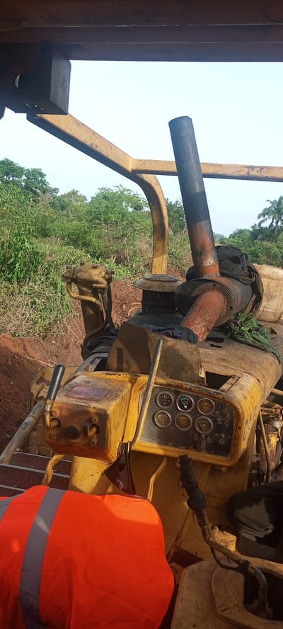 The image provides an eye-level view from within heavy yellow construction machinery, likely a bulldozer or track-type tractor, operating in an outdoor, rural setting in Otunade, Nigeria.

In the lower left foreground, a bright orange high-visibility safety vest with grey reflective stripes is draped over a component of the machine, suggesting operator presence or recent activity.

The midground is dominated by the well-used, weathered yellow machinery. A control panel is visible, featuring several circular analog gauges that appear to monitor functions like fuel, temperature, and pressure. Below these, various toggle switches are present, with visible labels including "HEAD LIGHTS," "START," "RESET," and "BRAKE." Multiple control levers with black grips are positioned around the panel, indicating operational controls for the machine. A prominent, rusty, dark-colored exhaust pipe rises vertically from the engine compartment, with dark fabric materials draped around its base. The robust yellow metal framework of the machine's roll-over protective structure (ROPS) or canopy extends above and around the operator's area.

The background showcases a vibrant, green landscape characterized by dense vegetation, including numerous trees and shrubs, consistent with a tropical environment. The ground immediately behind the machinery is reddish-brown earth, indicating recent disturbance or excavation. Further in the distance on the right, taller trees, possibly palm trees, dot the horizon. The sky is bright and clear, suggesting a daytime scene, possibly during the morning or late afternoon. The overall scene depicts heavy equipment at a worksite, likely involved in land clearing or earthmoving.