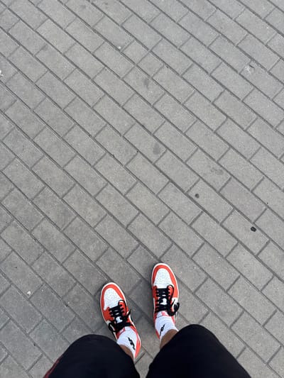 The image is an overhead shot of a person standing on a paved sidewalk. The sidewalk is made of rectangular gray bricks laid in a herringbone pattern. The person is wearing black shorts and white socks with black accents. On their feet are a pair of orange and white sneakers with black laces. The camera is angled downwards, capturing the feet and a portion of the person's legs. The lighting suggests it is daytime. The scene appears to be an outdoor urban environment, possibly a street or a public walkway. No specific landmarks of Białystok, Poland are visible. There is no text visible in the image.