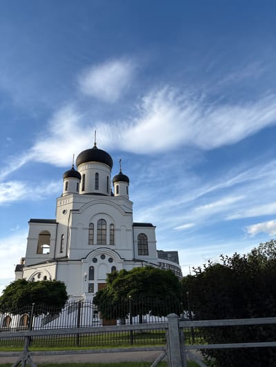 The image depicts a white Orthodox church with black domes, set against a bright blue sky filled with wispy white clouds. The church architecture is characterized by arched windows and multiple cupolas. The scene is bordered by a metal fence and lush green trees. To the right of the church, a modern, curved building is visible in the background. The lighting suggests it is daytime, with the sun casting shadows on the building and illuminating the sky. The overall atmosphere is serene and tranquil. No people are visible in the image, and no text is present. The orientation of the image is vertical, with the sky occupying the upper portion and the church and surrounding elements filling the lower part. The setting appears to be a public space, possibly a park or a church grounds, in Chelyuskinsky, Russia.