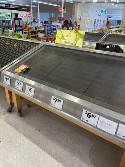 The scene is an empty checkout line in a grocery store in Middle Park, Australia. The checkout counter is made of metal and wood. The conveyor belt is empty except for a package of nuts sitting on the belt. The price tags for various items, including $7.90, $6.50, $6, $5.50, $4.50, and $5.50 are displayed in the frame. The floor is a speckled gray tile with a red rubber ball at the bottom of the frame. There is a white scale behind the checkout counter. In the background are red signs in an aisle, and a customer in a blue shirt can be seen in the distance.  The store is well lit with bright fluorescent lights.  The photo is taken from a low angle.
