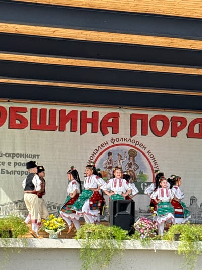 The image captures an outdoor performance on a stage set up in Pordim, Bulgaria. A group of children, dressed in traditional Bulgarian folk costumes, are dancing. The boys wear vests over white shirts and trousers with decorative trim, and black hats. The girls wear white blouses with embroidered details, colorful skirts or aprons, and headpieces adorned with flowers. They are positioned on a stage decorated with floral arrangements.
Behind the performers, a large banner displays red, stylized Cyrillic lettering, partially obscured by the dancers. A circular emblem with a depiction of traditional figures and the text "Национален фолклорен конкурс" (National Folklore Competition) is also visible on the banner. The stage is framed by a modern, multi-layered roof structure with dark grey and light brown wooden elements. The background behind the banner appears to be a mural or backdrop depicting buildings. The overall scene suggests a cultural event or festival taking place in the town.