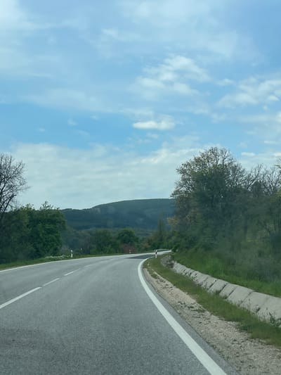 This image, captured in Madrets, Bulgaria, depicts a serene, winding asphalt road traversing a natural, mountainous landscape. The perspective suggests it was taken from a moving vehicle, looking ahead along the road.
In the foreground and mid-ground, the road features a clear solid white line marking its right edge and a dashed white line, possibly separating lanes or indicating a shoulder. To the immediate right of the road, a concrete curb or drainage channel runs alongside, bordered by green grass and light-colored earth. Lush green vegetation and a variety of trees line both sides of the road. Some trees exhibit sparse, dark branches, indicating either early spring or deciduous species, while others are full of vibrant green foliage.
In the background, a large, dark green mountain or hill rises majestically against the sky, its slopes densely covered in trees. The sky above is a bright, clear blue, adorned with scattered, fluffy white clouds, suggesting fair weather conditions. The natural lighting indicates it is daytime, likely morning or afternoon, given the soft, even illumination.
No people or visible text are present in the image, emphasizing the tranquil and undeveloped nature of the location. The scene evokes a sense of peaceful travel through a picturesque, rural or mountainous environment.