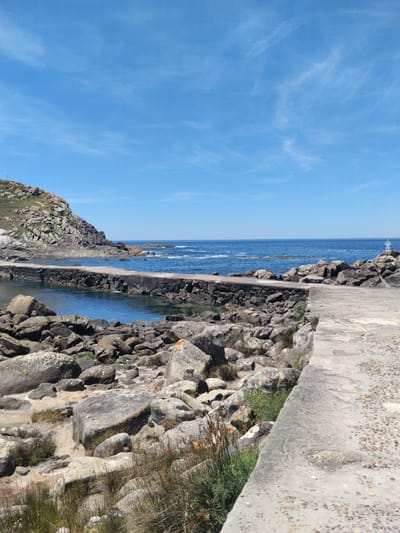 This is a scenic photograph of a coastal area in Vigo, Spain. The image features a clear blue sky with a few wispy clouds, extending over a calm blue ocean. In the foreground, a rocky shoreline is visible, with large, weathered boulders scattered along the edge. A concrete pathway or pier runs along the water's edge, with a stone wall bordering a sheltered inlet. The water in the inlet appears clear and shallow. Beyond the inlet, more rocky outcrops rise, and the ocean stretches out to the horizon. The overall impression is one of a natural, tranquil coastal landscape under a bright, sunny sky.