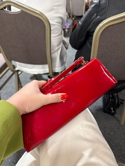 A person is holding a red, patent leather clutch bag. The person's fingernail is painted red. The person is wearing a green garment. The scene contains a chair with a grey seat and metal frame. A black handbag is visible in the background. White clothing is visible next to the clutch bag. The location is unknown.