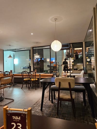 The image depicts the interior of a modern cafe. The perspective is from a seated position at a table, looking towards the entrance and through large glass windows into the street. The cafe is furnished with dark tables and chairs with some lighter upholstered seating. One prominent feature is a large, white, spherical pendant light hanging from the ceiling. Through the glass, several people are visible, some appearing to be staff members working behind a counter or in the kitchen area, and others possibly patrons. The floor is tiled with a grid pattern. In the foreground, a wooden stand with a sign displaying "TABLE" and the number "23" is visible. The overall atmosphere appears to be dimly lit, suggesting it is either evening or the cafe has ambient lighting. The presence of typical cafe furniture, lighting, and people engaged in what appears to be service or dining activities, positions this scene within an urban commercial establishment. This scene is not in Ngaglik, Indonesia; it is a cafe interior.