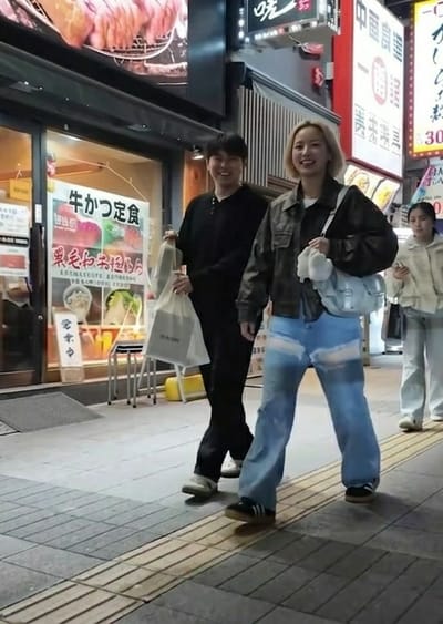 Three individuals are depicted walking from right to left along a paved sidewalk in a commercial district of Gifu, Japan, during low-light conditions. In the foreground, a young East Asian male, wearing a black long-sleeved Henley shirt and black trousers, walks while carrying two shopping bags—one transparent plastic, one white paper bearing "on the box" text—in his left hand. He exhibits a smile directed towards the viewer. Alongside him, a young East Asian female with short blonde hair wears a dark utility-style jacket, wide-leg light blue jeans with horizontal faded sections, and dark sneakers. She carries a light blue shoulder bag with a white fluffy accessory attached to its strap and is also smiling towards the viewer. Further back and to the right, a third young East Asian female, with dark hair and wearing a light-colored collared jacket and trousers, walks with a neutral expression.

To the left, a building facade features a large glass storefront window displaying Japanese promotional signs, prominently featuring "牛かつ定食" (Gyukatsu Teishoku – Beef Cutlet Set Meal) alongside images of food. Above this storefront, multiple illuminated Japanese signs are visible, including "暁一号店" (Akatsuki Store Number One) and a vertical sign for "中国料理" (Chinese Cuisine). The sidewalk consists of grey and light grey rectangular pavers and includes a yellow tactile paving strip for the visually impaired. The scene is illuminated by artificial light from the surrounding urban environment and establishments.