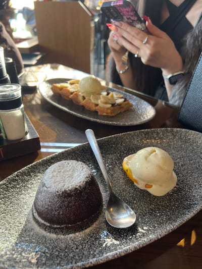 The scene depicts an indoor dining setting within a cafe or restaurant in Strumica, North Macedonia. Natural light illuminates the area from the left side of the frame. In the immediate foreground, a dark gray speckled rectangular plate contains a chocolate lava cake dusted with powdered sugar, a silver spoon, and a scoop of vanilla ice cream resting on a yellowish-orange half-fruit, consistent with a preserved peach or apricot. To the left of this plate, a clear salt shaker on a small wooden base is visible, alongside a dark, partially obscured object resembling a book or menu.
In the mid-ground, an oval dark gray speckled plate holds a waffle topped with sliced bananas, a dark drizzle (likely chocolate sauce), and another scoop of vanilla ice cream. Behind this plate, a person is partially visible, primarily their upper torso, head, and hands. This individual has long, dark hair and manicured hands with long fingernails painted in a red-to-orange ombre style. They wear multiple rings on their index, ring, and thumb fingers, a silver chain bracelet on the left wrist, and a white smartwatch on the right wrist. The person is actively holding and engaging with a dark-cased smartphone. The background is blurred, showing other tables and the general interior of the establishment.