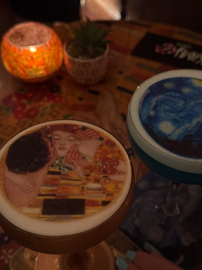 This media file captures a close-up view of two elaborately decorated beverage lids, likely for coffee or cocktails, resting on a table. The lid in the foreground features a reproduction of Gustav Klimt's famous painting "The Kiss," depicting a couple embracing amidst a richly patterned, gilded background. The lid in the background displays Vincent van Gogh's "The Starry Night," with its swirling blue sky and yellow stars.
Other objects visible include a small potted succulent in a textured pot and a decorative, illuminated candle holder with an orange, textured glass. The overall impression is one of an artistic and cozy setting, perhaps a café or a home environment. The lighting appears to be warm and ambient, suggesting it might be evening or a dimly lit interior. A person's hand with light blue nail polish is also visible on the right side of the frame, reaching towards one of the beverage lids.
While there is some text visible on a black label in the upper left portion of the image, it is blurred and illegible. The context provided, Valletta, Malta, suggests the photograph was taken in this location. The overall mood conveyed is one of appreciation for art and a moment of relaxation.