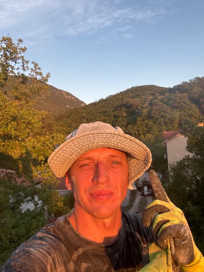 The media file is a selfie taken outdoors, likely in a rural or semi-rural area, given the presence of trees and a hillside. The main subject is a person wearing a light-colored hat and work gloves. The person's face is visible, and they appear to be looking at the camera. The person's right hand, covered in a yellow and black work glove, is extended and pointing towards the right side of the frame. The glove appears to be dusted with white particles, possibly plaster or paint. The person is also wearing dark clothing, which also has specks of the same white material.

In the background, there is a lush green hillside covered in trees. A building with a reddish-brown roof is partially visible amongst the trees. Further in the distance, a mountain or large hill is visible. The sky is clear and a pale blue, with a few wispy clouds. The lighting suggests it is either early morning or late afternoon, with the sun casting a warm glow on the scene. The overall mood appears to be casual and perhaps indicative of a work break or a moment captured during an outdoor activity. The location context provided places this scene in Prijeradi, Montenegro. No visible text is present in the image.