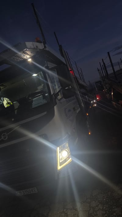 The image shows a Volvo car carrier truck parked on a cracked asphalt surface at dusk or dawn. The sky is a deep blue with some wispy clouds. The truck's headlights are on, casting bright beams of light. The truck has the "VOLVO" logo visible on the front. There is text on the side of the truck that includes "IVAS AUTO". A license plate is visible on the front of the truck, reading "CB 3150 KE". In the background, other vehicles and structures are dimly visible, with some red lights present. The overall scene suggests a commercial vehicle ready for transport or having just completed a journey. The location is Épila, Spain.
