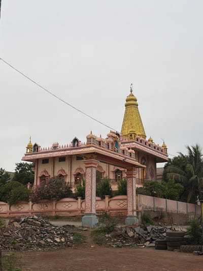 A temple, likely in Bugganipalli, India, is depicted under a cloudy sky. The temple features a prominent golden spire, decorative pink and cream colored facade with arched windows and ornate detailing. A covered entrance structure with decorative pillars stands in front of the main building. To the left of the temple, trees and foliage are visible, including a palm tree on the right. In the foreground, a pile of construction debris, including stones and tires, rests on the ground. A power line stretches across the upper portion of the image. The overall scene suggests an outdoor view of a religious structure with surrounding vegetation and signs of recent construction or demolition.