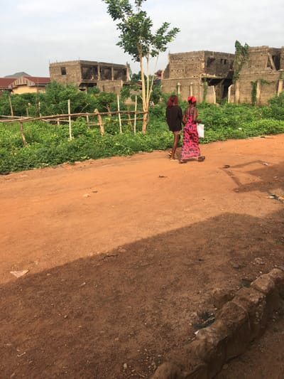 The image captures a scene in Isieke, Nigeria, showing two women walking on a dirt path alongside overgrown vegetation and unfinished buildings under an overcast sky.
**Subjects:** Two women are walking from left to right across the middle ground of the image. The woman in front wears a red patterned dress and a red head covering, carrying a white bag. Behind her, another woman is dressed in dark clothing, possibly a dark top and a dark skirt or wrapper, with dark hair or a dark head covering. Both appear to be wearing sandals. In the background, there are several large, multi-story concrete structures that appear to be either unfinished or derelict, with some green vines growing on them. To the far left, a tree with green leaves is visible.
**Scene and Setting:** The primary setting is a wide, reddish-brown dirt path or road that occupies the right half and foreground of the image. To the left of the path, there is a strip of dense green foliage, appearing wild and unkempt, with some wooden stakes or fences interspersed within it. Beyond the vegetation, the aforementioned concrete buildings rise. The sky, visible at the top left, is a uniform light gray, indicating an overcast day.
**Activity:** The two women are in the process of walking along the dirt path.
**Notable Details:** The time of day appears to be daytime, possibly mid-day, given the overall brightness, although the overcast sky diffuses direct sunlight. Shadows of unseen objects or individuals are cast on the dirt path. The buildings in the background, constructed from concrete blocks and lacking finishes, suggest a developing or economically challenged area. There is no visible text in the image. The environment, with its dirt roads, specific building styles, and vegetation, aligns with the provided location context of Isieke, Nigeria.