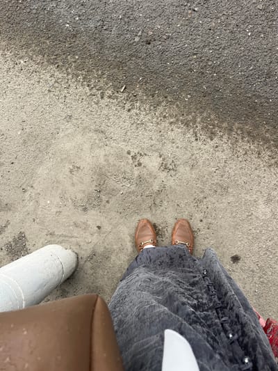 The image is an overhead shot of a person's lower body and feet, standing on a wet, asphalt and concrete surface. The person is wearing brown, horse-bit loafer-style shoes and a dark gray, flowing skirt with visible button detailing. To the left of the person is a white, cylindrical object, possibly a bollard or pipe, and a brown bag or seat. The ground appears to be a pathway or roadside, with patches of darker, wet asphalt transitioning to lighter, damp concrete. There are small stones and debris visible on the ground. The overall environment suggests an outdoor urban setting, possibly in Medrese, Kyrgyzstan, given the context. There are no other people or animals visible in the frame. The action depicted is standing still.