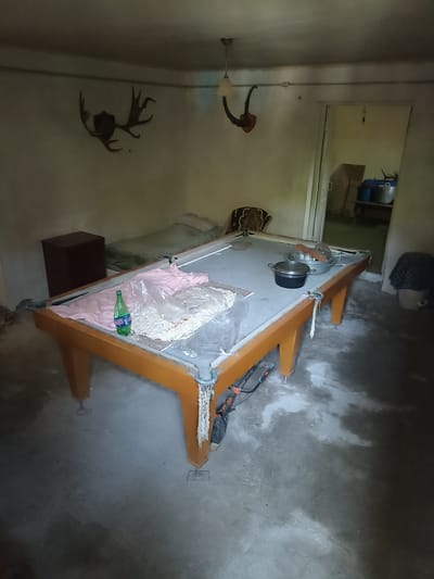 The image depicts an indoor scene in the city of Aygezard, Armenia. The room contains a pool table with orange legs and a grey playing surface. On the pool table, there is a pink cloth, a green bottle of what appears to be soda, and a plastic bag filled with small white objects. There are also two pots and a wooden item on the table.

To the left of the pool table, there is a dark brown cabinet and a bed with a green cover and a light-colored pillow. The walls are a pale yellow color. Mounted on the wall above the bed are two sets of animal antlers. A light fixture hangs from the ceiling.

In the background, a doorway leads to another room. Through the doorway, several blue containers and metal pots are visible. A bucket with a dark patterned cloth is situated to the right of the pool table. The floor appears to be concrete and has some dusty or wet patches. The overall atmosphere suggests a somewhat neglected or sparsely furnished space.