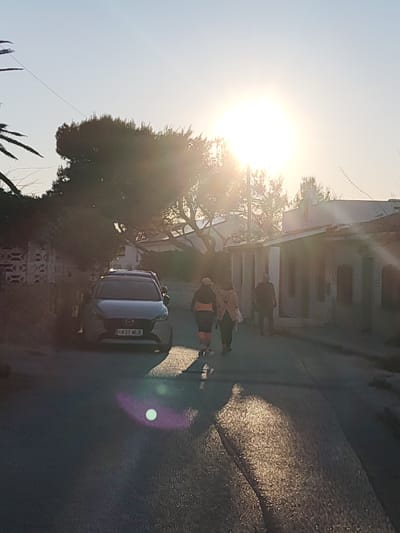 This image depicts a sun-drenched street scene in Rotes, Spain, likely captured during the early morning or late afternoon due to the low sun angle and prominent backlighting. The bright, clear sun dominates the upper center-right, casting strong light and creating significant lens flare, including a distinct circular purple and white orb near the bottom-center of the frame.
Three individuals are walking away from the camera down the asphalt road, their forms largely silhouetted against the bright light. Two are walking together on the left side of the road, with one wearing an orange band visible around their lower torso. A third person is further ahead on the right side of the street.
A dark grey Mazda car, identifiable by its model characteristics and visible emblem, is parked on the left side of the road, facing away from the viewer. Its Spanish license plate, "6831 MLD," is clearly legible. The street is lined with mature trees, including a palm tree on the far left and other lush green foliage that partially obscures the houses. Residential buildings with light-colored walls are visible on both sides of the street, some appearing to be single-story structures. The sky above is a clear, pale blue. The road surface exhibits textured patches where sunlight reflects intensely. The scene conveys a tranquil, sunny atmosphere in a residential area.
