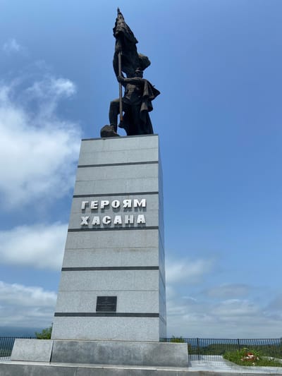 The image displays a monument in Kraskino, Russia, dedicated to the "Heroes of Khasan." A dark bronze statue of a figure holding a flag stands atop a tall, grey granite obelisk. The obelisk features the inscription "ГЕРОЯМ ХАСАНА" (To the Heroes of Khasan) in prominent silver letters. A smaller plaque is affixed to the lower section of the obelisk. The monument is situated outdoors, with a clear blue sky and scattered white clouds forming the background. The surrounding landscape, visible in the distance, appears to be hilly and green, bordered by a black metal fence. A small floral arrangement rests near the base of the monument.