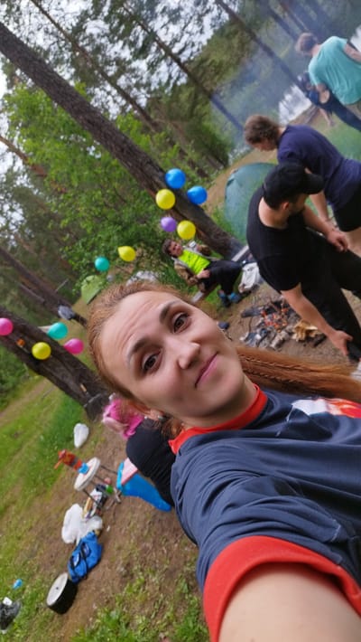The image depicts an outdoor gathering in a wooded area, possibly a park or campsite. A woman in the foreground is taking a selfie, smiling at the camera. She has long, reddish-brown braided hair and is wearing a navy blue shirt with red trim. In the background, several other individuals are visible, some sitting and others standing. One person is wearing a bright green jacket, another a dark blue shirt, and another a light blue shirt. There are several colorful balloons scattered around the scene, some tied to trees. Tents and camping equipment are also present, suggesting a camping or outdoor event. A small campfire is visible in the middle ground, with smoke rising from it. The ground is covered with grass and dirt, and tall trees with green foliage dominate the background. The overall atmosphere appears relaxed and casual. The location is identified as Orekhovo, Russia.