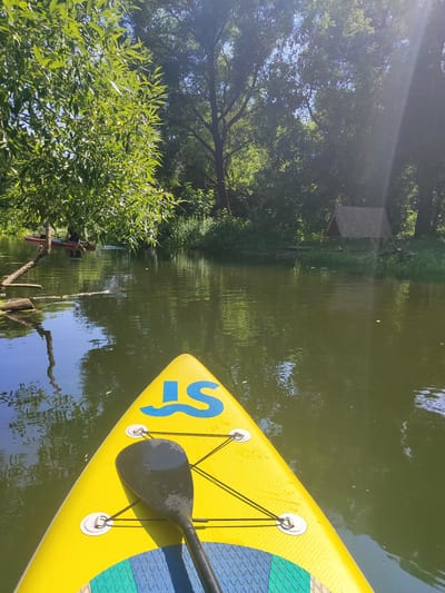 The image captures a serene scene on a body of water, likely a river or canal, in Bol'shoye Alekseyevskoye, Russia. The perspective is from a yellow stand-up paddleboard (SUP), with a black paddle resting on its surface. The board features blue lettering that appears to be "JS". In the foreground, the water is calm and reflects the lush green foliage that lines the banks. Further down the waterway, other individuals are visible on red paddleboards, also enjoying the tranquil environment. The background is dominated by dense trees, with sunlight filtering through the leaves, creating a dappled effect. A small, rustic wooden structure with a pointed roof is situated on the right bank, adding to the natural and somewhat secluded atmosphere of the location. The overall impression is one of peaceful outdoor recreation on a clear, sunny day.