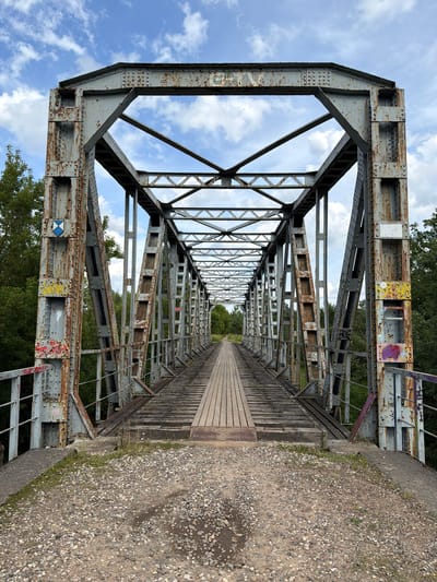 An overcast day with partly cloudy skies looms over a decaying metal truss bridge in Tēraudi, Latvia. The bridge, constructed from rusted steel beams and rivets, is supported by two large, weathered support towers. A wooden walkway, showing signs of wear and tear, spans the length of the bridge, flanked by a lower level of older wooden planks. Graffiti is visible on the bridge's support structures. Dense green trees surround the bridge, hinting at a natural or semi-rural setting. The ground leading to the bridge is covered in gravel and dirt, with some patchy grass. The overall scene suggests neglect and disuse.