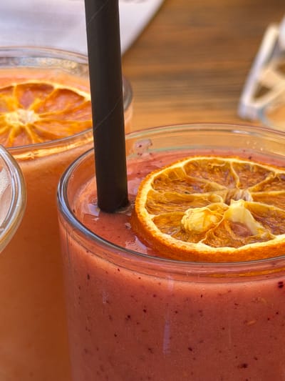 The image presents a close-up, high-angle shot of two glasses filled with smoothies, likely taken in Cala D'or, Spain. The smoothies have a reddish-brown hue with visible specks, suggesting a berry-based recipe. Each glass is garnished with a dried orange slice clinging to the rim. A black straw pierces the smoothie in the foreground glass. The background reveals a wooden surface and the edge of another glass object. The composition is slightly tilted, adding a dynamic feel to the scene.