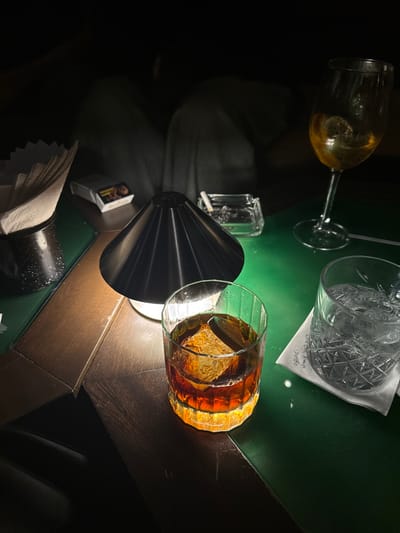 A close-up, low-light image depicts a table scene at ONG AHAU DIVINO in Guadalajara, Mexico. The table features a split surface, with dark wooden sections on the left and a reflective, deep green surface on the right. A central black, conical table lamp emits a warm light. In the immediate foreground, a ribbed glass tumbler holds a dark amber beverage with a single large, square ice cube. To its right, a cut-glass tumbler contains clear liquid and ice, resting on a white coaster with the partial inscription "ONG AHAU DIVINO". In the upper right, a stemmed wine glass holds a light amber liquid with ice. To the left of the lamp, a dark container holds white paper napkins, positioned near a white pack of Marlboro cigarettes. Behind the lamp, a clear glass ashtray contains ash and a lit cigarette. Faintly visible in the blurred background are the light-colored pants of a person seated at the table.