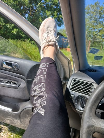 The image shows a person sitting in the driver's seat of a car. The person is wearing black Reebok leggings and a light-colored sneaker, which is placed on the open car door frame. The car's interior is visible, including the dashboard, steering wheel, air vents, and door controls. The outside environment is visible through the open door and windshield, showing trees and grass. There is no other information that specifies any location in the city of Sokolovo, Bulgaria.