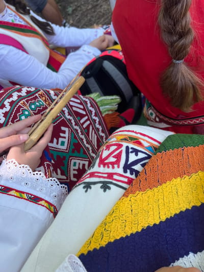 A close-up, high-angle view captures several individuals adorned in vibrant traditional Bulgarian folk attire during an outdoor gathering in Bolyarovo, Bulgaria. In the left foreground, a person's hands are clasped around a light-colored wooden wind instrument, resembling a kaval, which rests across their lap. This individual wears a white blouse with intricate eyelet lace trim on the sleeve cuff, below which is a band of colorful geometric embroidery in red, yellow, and green. Their lap is covered by a richly embroidered maroon or deep red fabric displaying prominent white and green stylized geometric patterns. To the right, another person is visible, wearing a plain red garment, from which a thick, dark brown braided ponytail, secured with a light gray hair tie, extends downwards. Adjacent to this individual and partially overlapping the embroidered fabric is a textured, horizontally striped woven item in bold bands of green, orange, yellow, and dark blue. In the upper left background, parts of another person's white garment with subtle embroidery are visible. The ground beneath the participants is indistinct, suggesting a natural, earthy surface under dappled light.