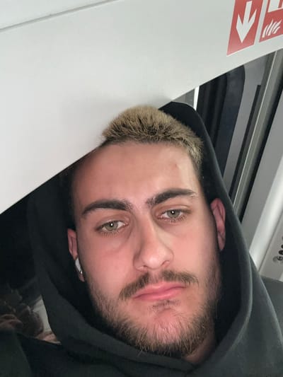 A close-up, rotated portrait shows a young man with striking green/hazel eyes, looking directly at the camera. He has short, light-blonde hair on top and darker stubble on his chin and cheeks. He is wearing a black hoodie, which frames his face, and a white earbud is visible in his left ear. The man appears to be resting his head against a light-colored surface, possibly a seat or wall.
The background suggests an interior setting, likely public transport such as a train or tram, given the overhead paneling and a dark, possibly windowed area behind him. To the left, a white panel features a red sign with a white arrow pointing right, accompanied by a symbol resembling stylized flames or smoke. This is indicative of a safety or emergency exit instruction, commonly found in transport vehicles. The overall lighting is soft but clear, suggesting it could be daytime inside the vehicle. The man's expression is neutral, bordering on pensive. Given the context, the setting is likely within a train or tram in Leverkusen, Germany.