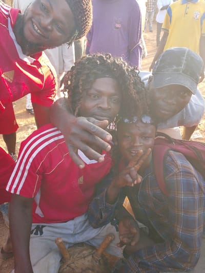 Four dark-skinned men pose closely together. One man with curly hair embraces another wearing a red and white Adidas shirt. A third man in a baseball cap leans in from the side. At the front, a young man in a plaid shirt flashes a peace sign. They are likely outdoors, based on the lighting and background showing other people. A small drum is visible at the bottom center.