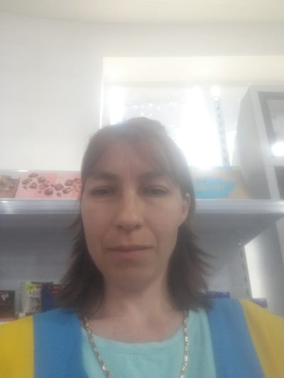 The image appears to be a selfie taken inside a store in Бородулиха ауылдық округі, Kazakhstan. A woman with brown hair is facing the camera. She is wearing a light blue shirt with yellow and blue stripes on the left side. A silver necklace is visible around her neck. Behind her, a shelf with various products is partially visible, including some chocolates in heart-shaped wrappers. There is a bright window and a white wall in the background. The lighting is somewhat harsh, with glare from the window. There is also a gray shelf with a box of cookies or treats with blue writing on the front.