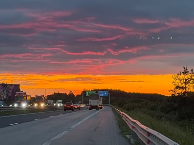 The image depicts a highway scene during a vibrant sunset. The sky is a dramatic canvas of orange, pink, and purple clouds, with streaks of lighter hues stretching across the twilight. A line of bright lights, possibly from a satellite train or aerial phenomenon, is visible in the upper portion of the sky. The highway itself is well-lit by the headlights of various vehicles, including several trucks and a red car. The road appears to be a multi-lane highway with clear lane markings and a metal guardrail on the right side. Lush green trees and vegetation line the sides of the highway, with a cluster of trees silhouetted against the bright sunset on the right. Road signs are visible, indicating directions. The overall impression is one of travel and movement against a breathtaking natural backdrop. This scene is located on the A1 highway, specifically near the exit for Baliovtsi, Bulgaria.