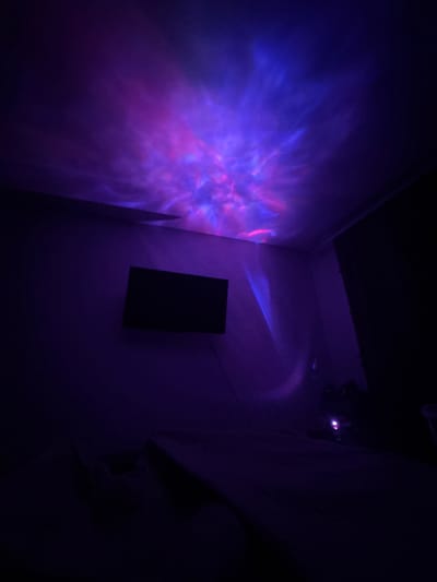 In a dimly lit room, a ceiling projector casts a vibrant nebula pattern of purples and blues onto the ceiling and upper walls. A black flat-screen television is mounted on the wall, facing a bed draped in dark fabric. The room appears to be a bedroom or a relaxation space. The scene is devoid of people, and the primary focus is on the dynamic light display and the sparse furnishings. The location is specified as Zhigulyovsk, Russia.