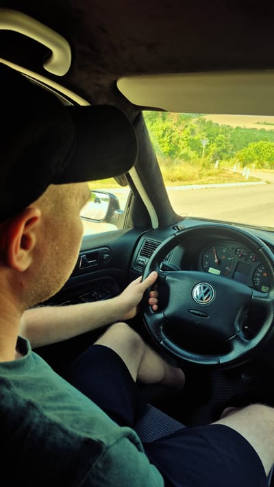 A man wearing a black baseball cap and a green t-shirt is driving a Volkswagen car on a sunny day. The interior of the car is visible, including the dashboard with its instruments, the steering wheel, and the door panel. The driver's hands are on the steering wheel, and his legs are visible through the open-sided shorts he is wearing. Outside the car, a rural landscape with green trees and fields can be seen through the windows. The man is looking ahead, presumably at the road, with another car visible in the rearview mirror. The overall scene suggests a journey through the countryside. It is unlikely this image was taken in Mihailovca, Moldova, as the car appears to be driving on the right side of the road, and Moldova drives on the right.