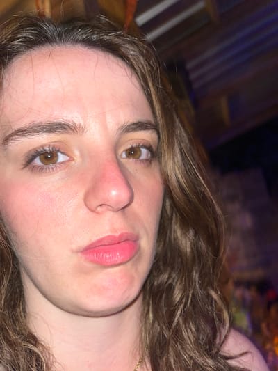 A close-up, slightly angled, indoor shot shows a young woman's face. She has brown hair, light skin, and brown eyes. Her lips are pink and appear to have lip gloss or lipstick on them. There is a slight red coloration on her cheeks and nose. The background is dark and indistinct, with some blurred lights visible. No specific location information is discernible from the image.