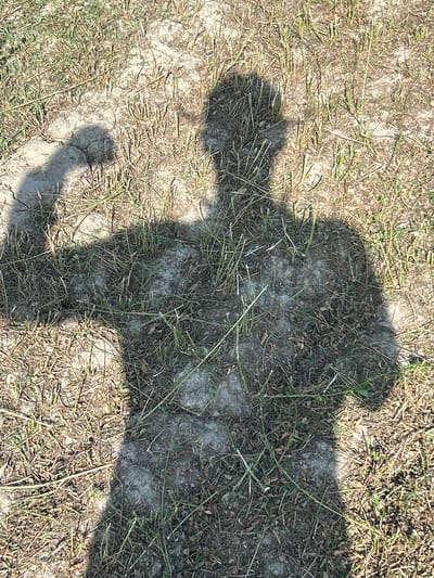 A shadow of a person is cast on dry, cracked earth with sparse, dry grass. The shadow is in a flexing pose, with one arm raised and bent at the elbow. The head of the shadow is indistinct. The scene is outdoors, likely in a rural or unpaved area, and the lighting suggests it is daytime. The location is specified as Kasri Arifan, Uzbekistan.