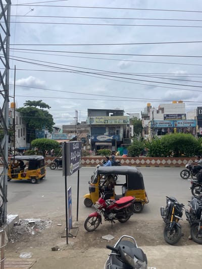 A street scene in Kurnool, India features a traffic intersection with several auto-rickshaws and motorcycles. In the foreground, a yellow auto-rickshaw is parked on the left, and a red motorcycle is positioned in the center, facing right. To the right, two black motorcycles are parked. In the background, other vehicles and pedestrians are visible, along with buildings lining the street. The sky is overcast with clouds.