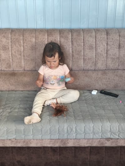 A young girl sits on a sofa with a light blue, paneled wall behind her. The sofa is upholstered in a taupe fabric with vertical channel tufting. The girl is wearing a light pink t-shirt with a "Happy Birthday" graphic featuring cupcakes and balloons, and cream-colored pants with white socks. Her legs are crossed, and she is holding a small blue object in her hands, looking down at it. To her right, on the sofa, are a white wireless earbud case and a black TV remote control. A brown doll wig is on the sofa near her left foot. The scene appears to be indoors, possibly in a living room or playroom. The lighting suggests it is daytime.