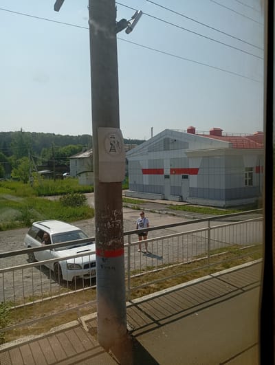 The image captures a street scene in Vostochnaya, Russia, on a sunny day. In the foreground, a concrete utility pole with a visible sign displaying symbols of a train and pedestrian figures stands to the right. The pole is weathered and has peeling paint, with a red band visible near the bottom. To the left of the pole, a white station wagon is parked. The car's driver, a woman with blonde hair wearing sunglasses, is looking out the window.
Behind the car and the utility pole, a metal fence separates the road from a paved area with scattered grass. Further back, a woman in a white t-shirt and dark shorts stands in the paved area, casting a shadow on the ground. A modern building with a red roof and grey and white striped facade is visible behind her, featuring several windows and doors. The background consists of lush green trees and a clear blue sky. The overall impression is of a utilitarian area adjacent to a building and residential or undeveloped land.