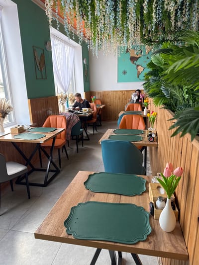 The image depicts an interior view of a restaurant. The restaurant is decorated with plants, including hanging floral arrangements and potted greenery. The tables are wooden with place settings consisting of green placemats. One table features a vase with pink tulips. Seating includes a mix of blue and orange chairs. Several people are seated at tables in the background, some engaged in eating or other activities. A world map is displayed on the wall. The restaurant appears to be located in Semenovka, Kyrgyzstan.