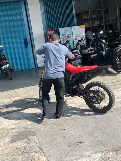 A man in a grey t-shirt and black pants stands on a concrete surface next to a red and black dirt bike, facing away from the camera. The man is pushing the dirt bike. In the background, there is a building with large blue roll-up doors and a white wall. Several other motorcycles are parked in the background, and posters are displayed on the wall of the building. The ground is cracked and weathered concrete, with shadows from the motorcycles and the man cast across it. The overall scene suggests an outdoor area, possibly a motorcycle repair shop or a parking area, in a sunny environment. The license plate on the dirt bike reads "W 5317 WBN". The location is Gedang, Indonesia.