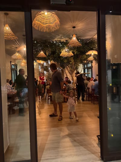 The image captures the interior of a restaurant in Punta Sam, Mexico, viewed through a glass entrance. The restaurant is decorated with numerous woven rattan pendant lights, casting warm shadows on the wooden floor and walls. Several patrons are seated at tables, some are walking, and staff members in uniforms are visible, suggesting a busy dining environment. In the foreground, a man in shorts and sandals stands near a young girl in a light-colored dress. Their figures are partially reflected in the glass, adding to the depth of the scene. The overall ambiance appears to be relaxed and inviting, with a mix of natural elements and modern design.