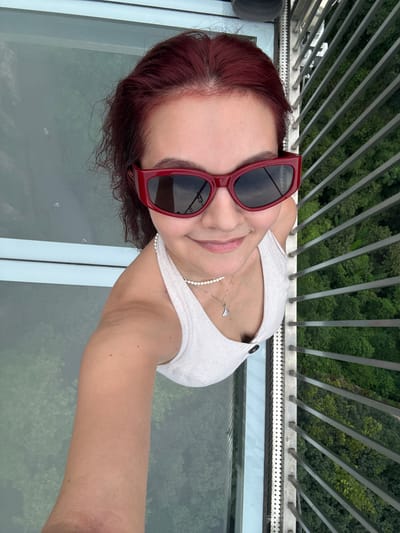 A young woman with red hair and red-rimmed sunglasses is taking a selfie from a high vantage point, possibly a glass-floored observation deck. She is wearing a white tank top and a pearl necklace with a pendant. To her right, a metal railing separates her from a dense, green forest below. The woman is smiling and looking at the camera. The image appears to be taken at the Qingyuan Sky Tower in Qingyuan City, China, known for its glass-bottomed observation deck offering panoramic views of the surrounding landscape.