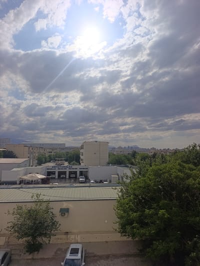 The image depicts a city scene in Ashgabat, Turkmenistan, under a partly cloudy sky with bright sunlight filtering through. In the foreground, a low-rise building with a beige facade and a green corrugated metal roof is visible. An air conditioning unit is mounted on the wall of this building. To the right, a large, dense green tree dominates the frame.
Further back, a commercial building with a sign reading "AWTOYUWALGA" (likely a car wash or auto service) is prominently featured. Several cars are visible within or near this building, suggesting an active car service operation. A white tent-like structure is also present in front of this building, possibly an outdoor seating area. Beyond this, a series of multi-story buildings, characteristic of urban development, are seen, with distant mountains or hills forming the horizon. The overall impression is of an everyday urban environment in Ashgabat, with commercial activity and residential buildings.