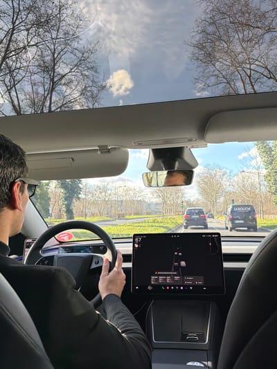 The image presents an interior view from the passenger seat of a modern vehicle, specifically a car featuring a yoke-style steering wheel and large central touchscreen, typical of certain electric vehicle manufacturers. The scene is set on a road in Villeurbanne, France, under a partially cloudy blue sky with visible sun glare reflected in the panoramic glass roof.

A male individual is observed from behind in the driver's seat. He has dark hair, wears dark sunglasses, and is dressed in a dark long-sleeved garment. His right hand is positioned on the black yoke-style steering wheel, which has a small circular red sticker affixed to it. A digital display behind the steering wheel shows the number "67," likely indicating speed.

Centrally located on the dashboard is a large horizontal touchscreen display. It actively displays a navigation interface with a 3D representation of the car's current position and surrounding traffic. The number "67" is also visible at the top-left of this screen, alongside a temperature reading of "20.0 C" and various application icons at the bottom. Overhead, a console houses a rearview mirror, reflecting the top of the driver's head, and a sensor unit. Sun visors are stowed in their upward position.

Outside the vehicle, the car is positioned on a multi-lane asphalt road. Directly ahead, two vehicles are visible: a dark SUV and a dark commercial van. The rear of the van prominently features the white text "LA BOUCLE." In the distance, a traffic light is illuminated green. The roadside and median are characterized by green grassy areas and numerous deciduous trees, most of which are bare, suggesting a colder season. Distant low-rise urban structures are faintly discernible through the trees.