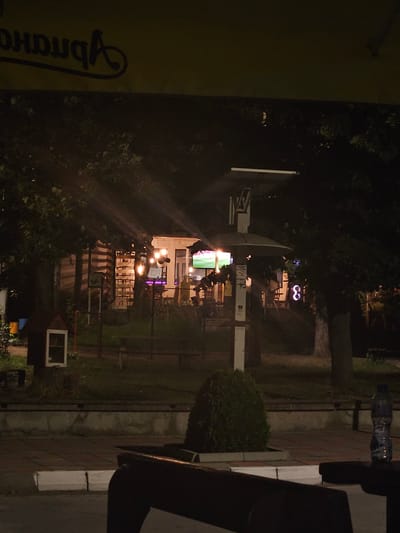 The image captures a nighttime scene in the city of Byaga, Bulgaria. In the foreground, dark silhouettes of outdoor seating, likely a table and chairs, are visible. The middle ground reveals a park-like setting with trees and scattered lighting. A small, red, birdhouse-like structure with a white window is situated on the left side of the frame. Beyond this, a building with illuminated windows suggests an indoor space. Several string lights are visible, casting a warm glow. A modern lamp post with a flat canopy stands in the center. In the background, trees obscure much of the view, but a bright screen displaying an image is visible through the foliage. On the right, a stylized illuminated number "8" is affixed to a tree. The overall ambiance is that of a relaxed outdoor establishment at night.